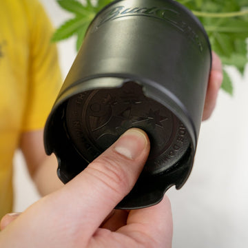 Finger pushing the bottom plate of the black BudCups' easy transplant mechanism with a blurry cannabis plant on the background