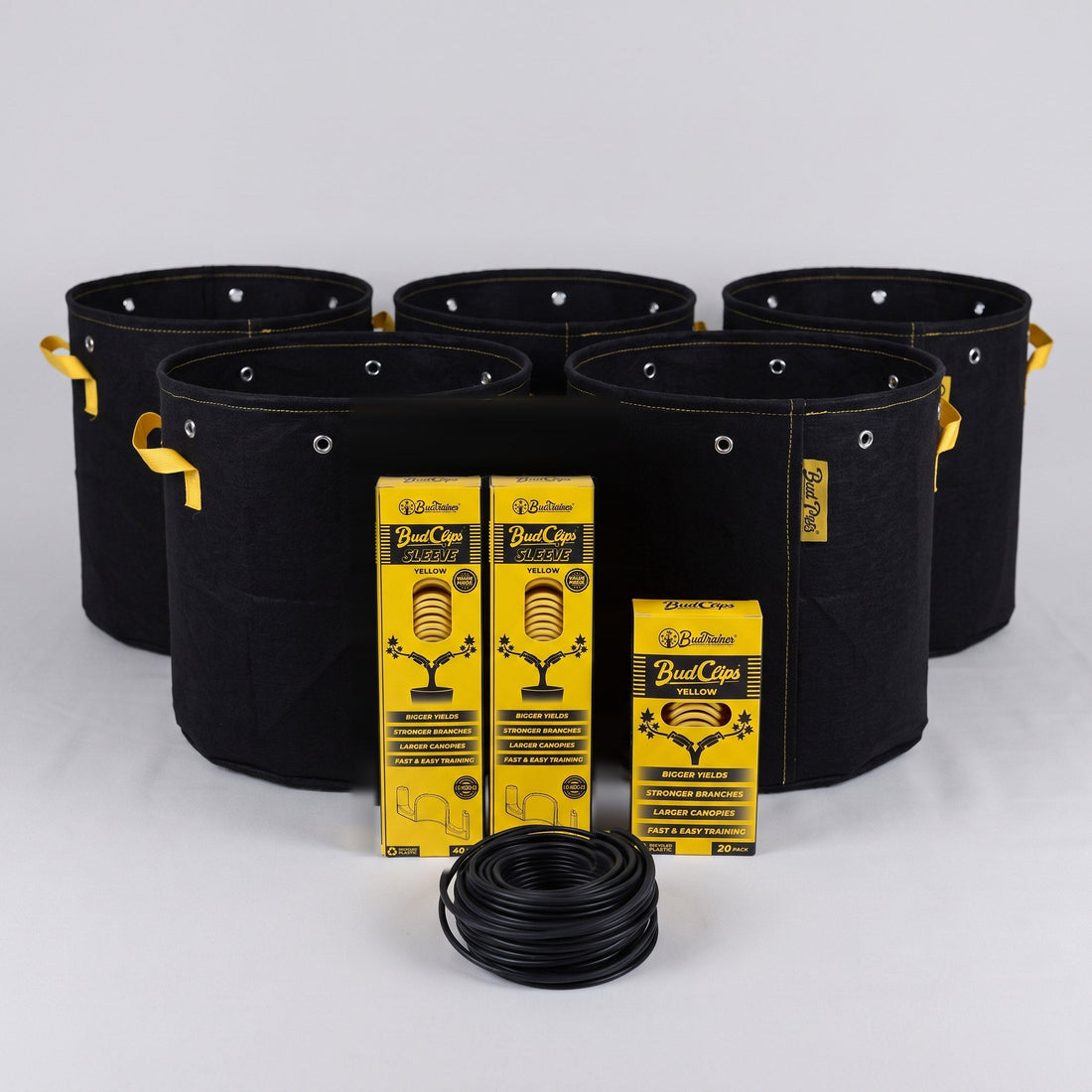 Five black BudPots fabric grow bags with two yellow BudClips 40-pack sleeves, a 20-pack box and a coil of black BudHuggers garden wire in front, on a plain grey background.