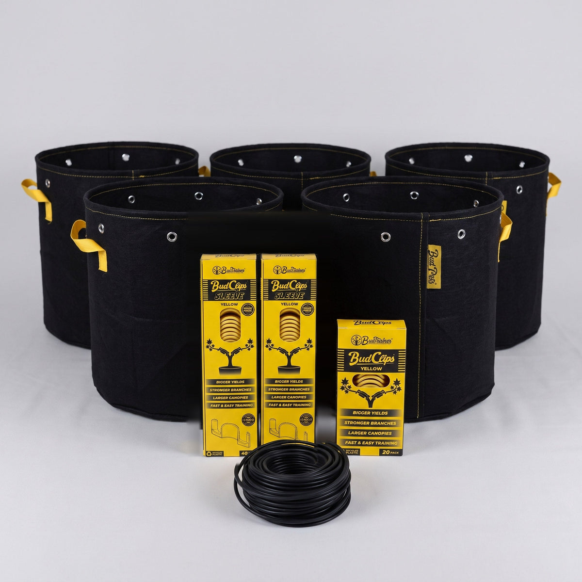 Five black BudPots fabric grow bags with two yellow BudClips 40-pack sleeves, a 20-pack box and a coil of black BudHuggers garden wire in front, on a plain grey background.