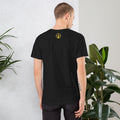 Man wearing a black t-shirt with a yellow logo on the back, standing against a light gray wall with plants.