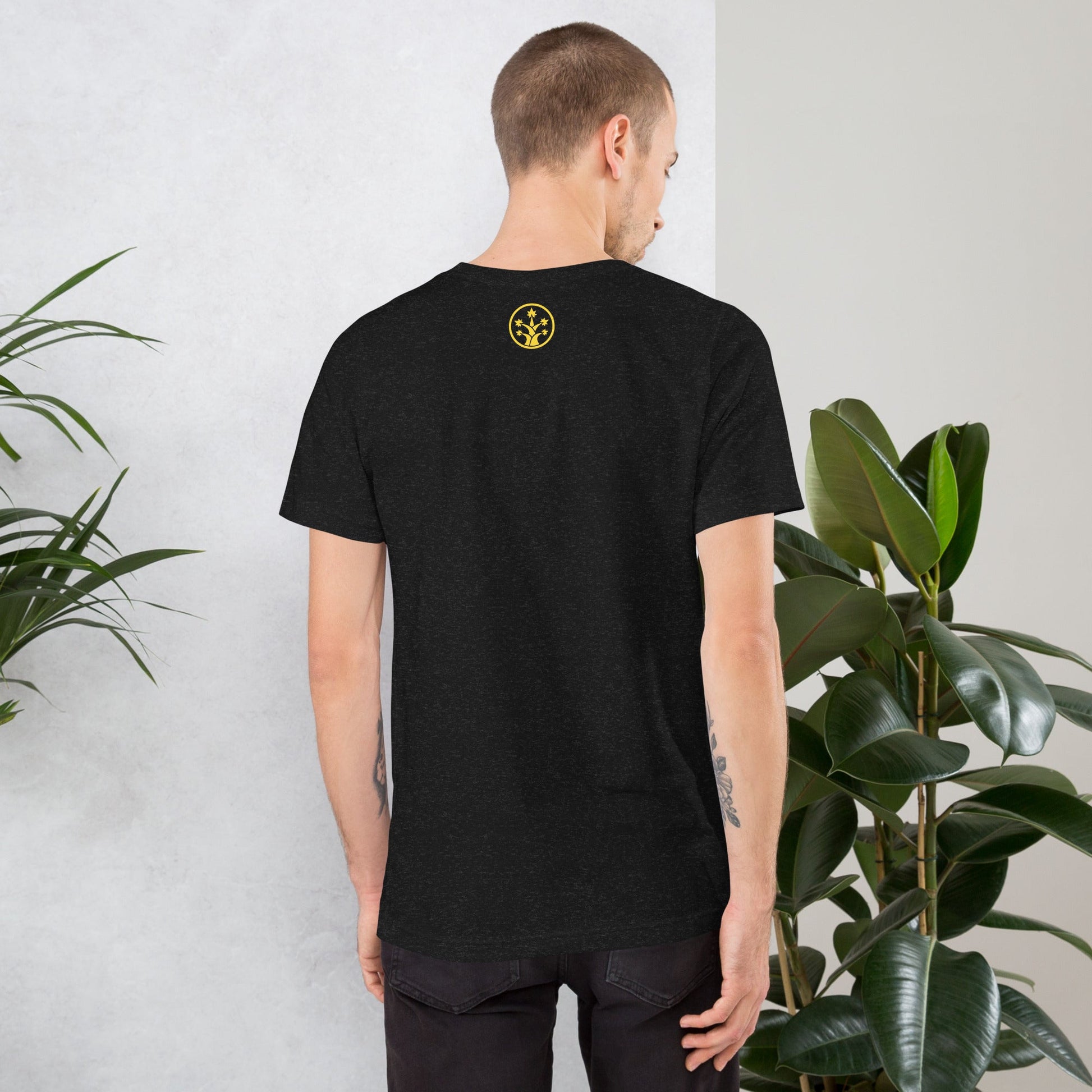 Man wearing a black t-shirt with a yellow logo on the back, standing against a light gray wall with plants.