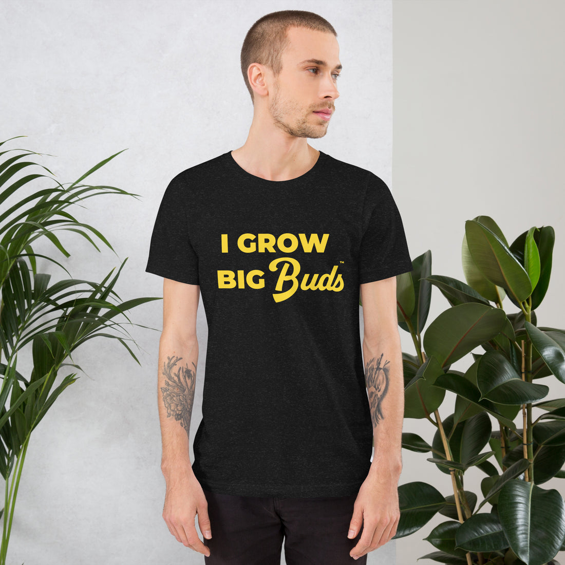 Man wearing a black BudTrainer t-shirt with 'I Grow Big Buds' in yellow text, standing against a white wall with plants.