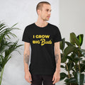 Man wearing a black BudTrainer t-shirt with 'I Grow Big Buds' in yellow text, standing against a white wall with plants.