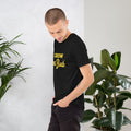 Man wearing a black BudTrainer -shirt with yellow text standing next to green plants.