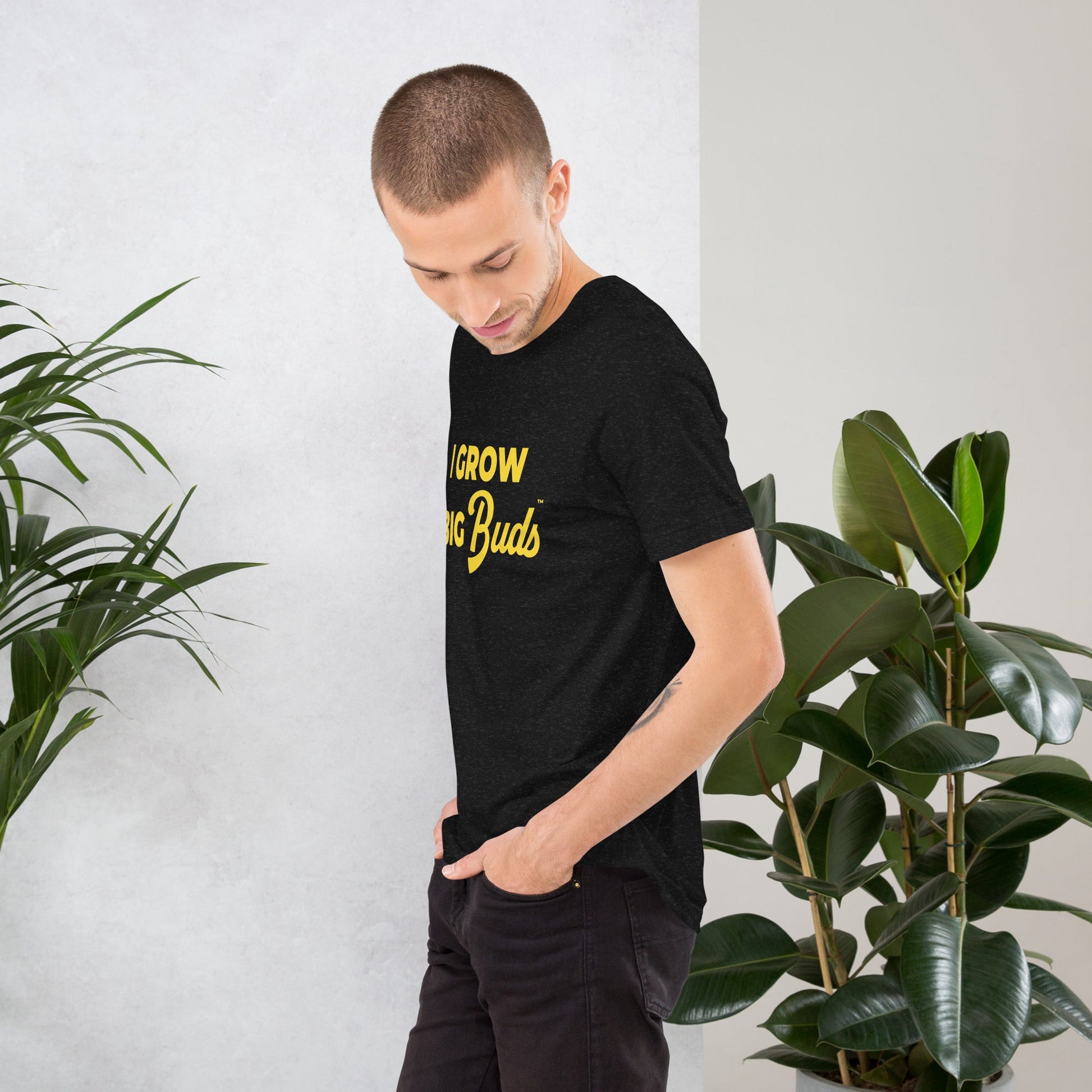 Man wearing a black BudTrainer -shirt with yellow text standing next to green plants.