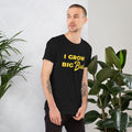 Man wearing a black BudTrainer t-shirt with 'I Grow Big Buds' text, standing between two potted plants.