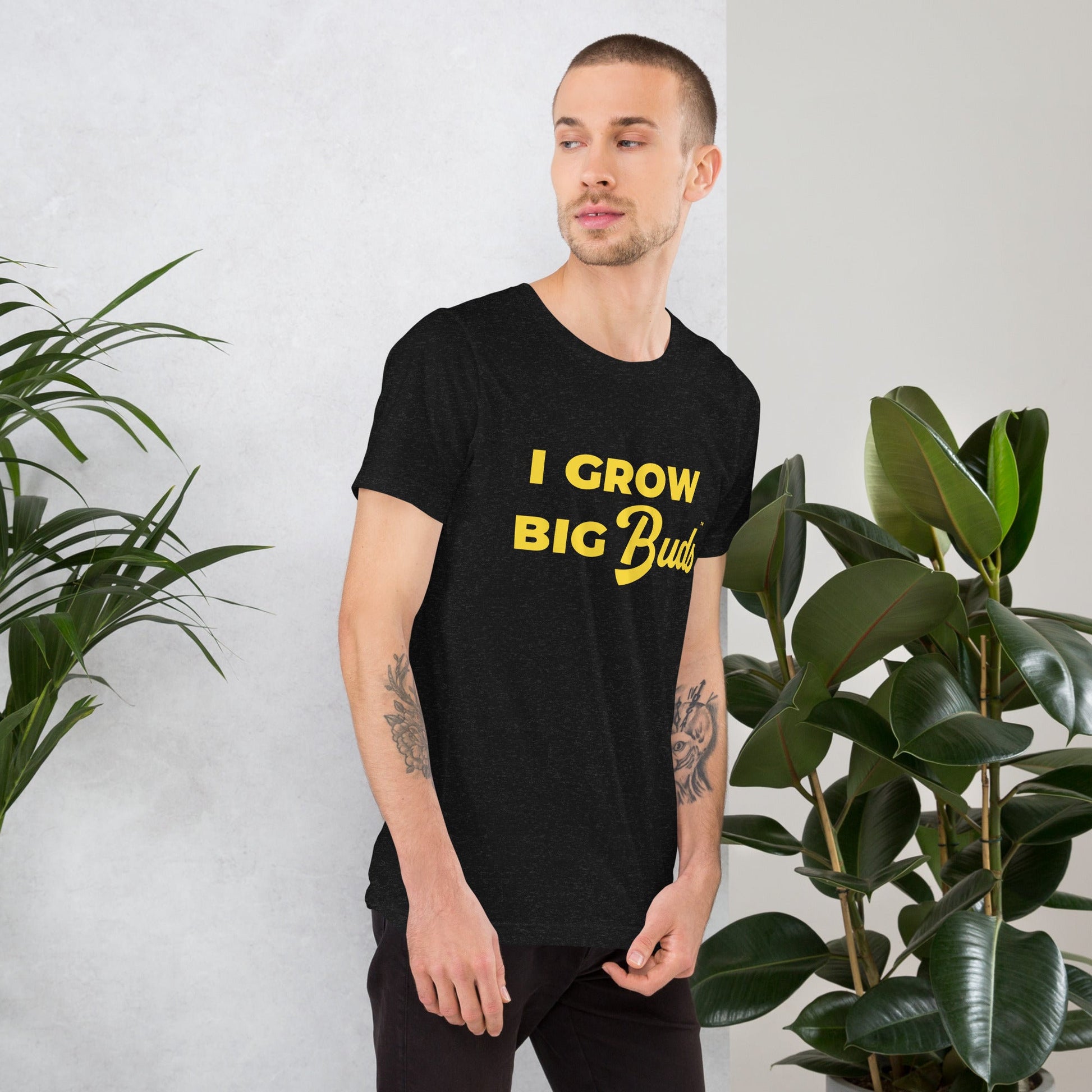Man wearing a black BudTrainer t-shirt with 'I Grow Big Buds' text, standing between two potted plants.