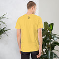 Man wearing a yellow BudTrainer t-shirt with a logo on the back, standing against a light gray wall with plants.