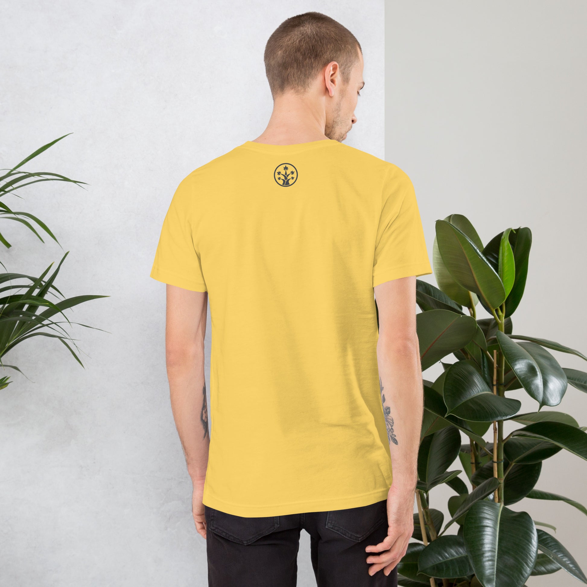 Man wearing a yellow BudTrainer t-shirt with a logo on the back, standing against a light gray wall with plants.