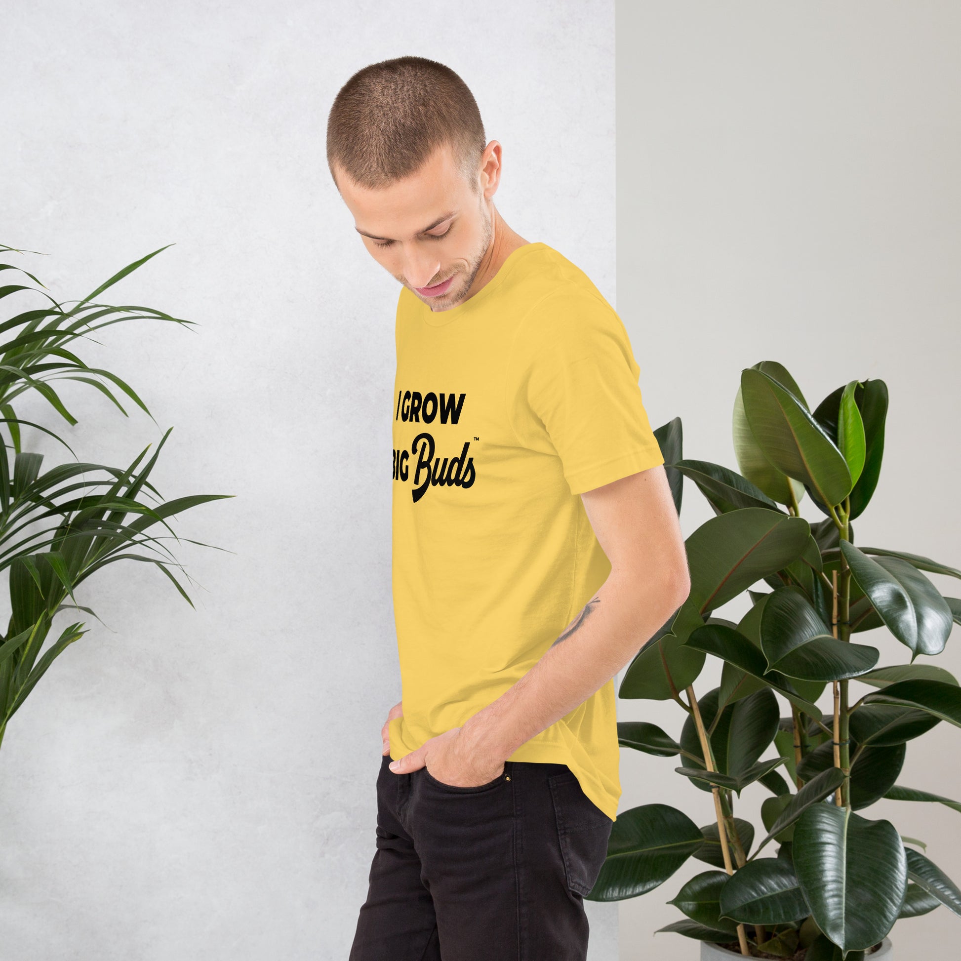 Man wearing a yellow BudTrainer t-shirt with text standing between two potted plants.