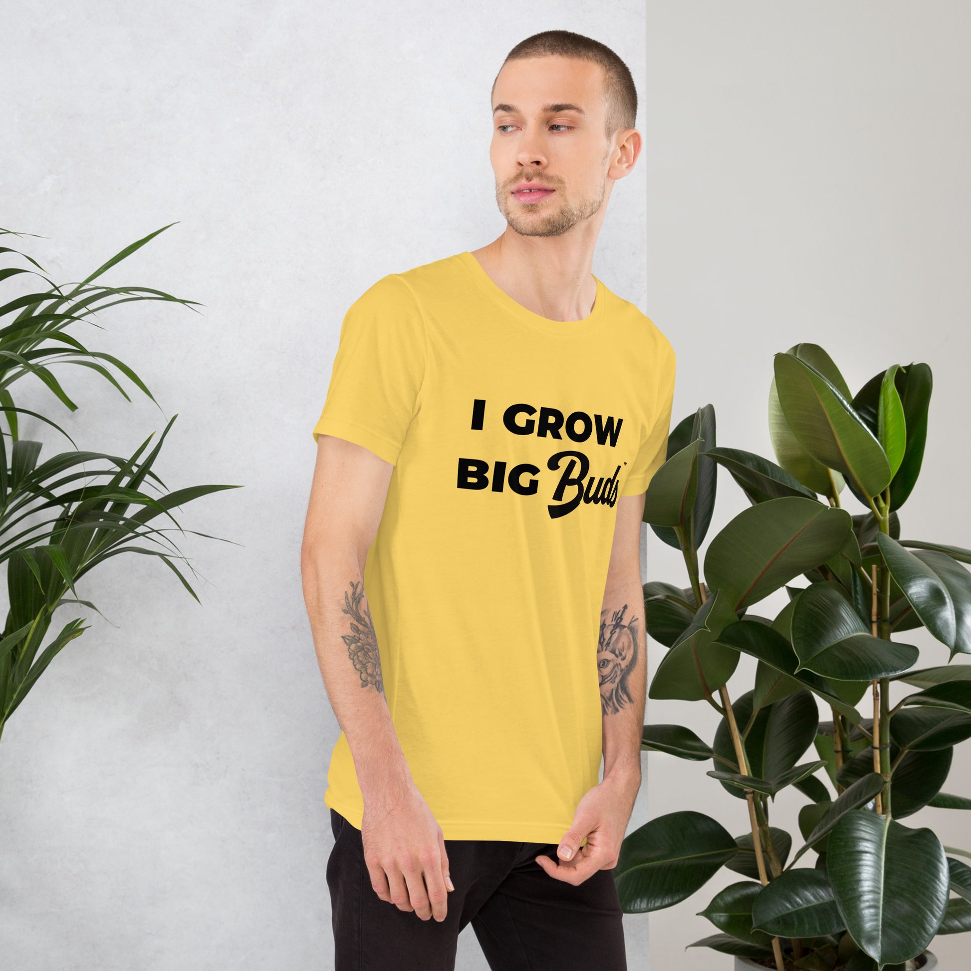 Man wearing a yellow BudTrainer t-shirt with 'I Grow Big Buds' text, standing between two potted plants.
